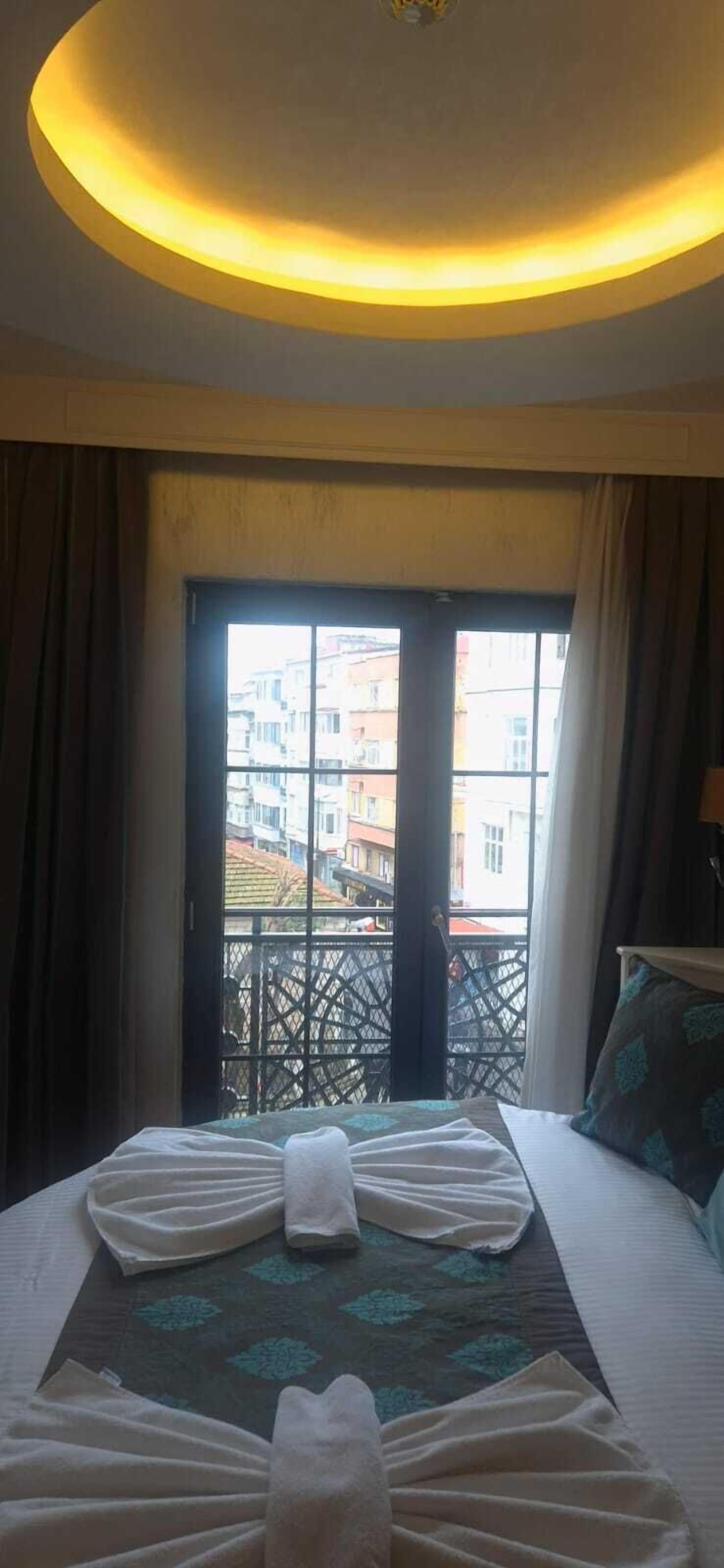 A hotel room with a bed, a window with a view of buildings, and a ceiling light.