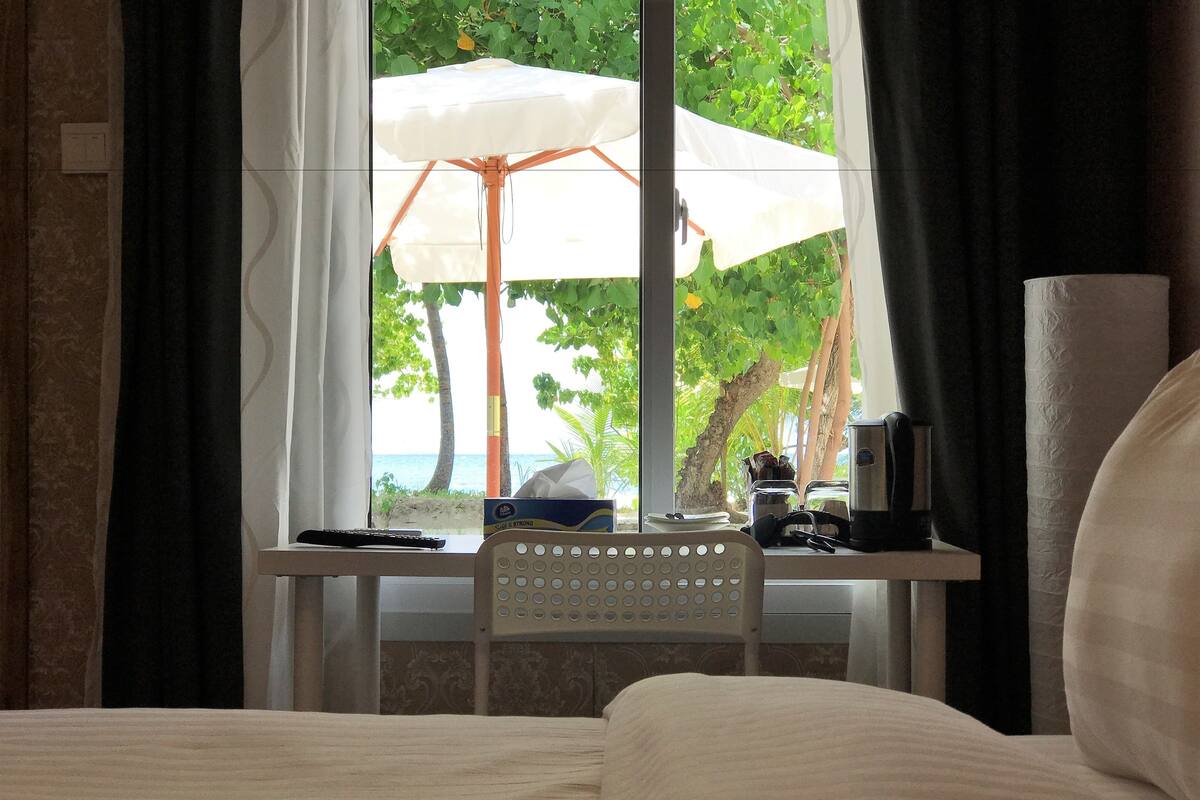 A hotel room with a bed, a small table with a remote control, a coffee maker, and a view of the beach through a window with curtains.