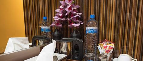 Una recepción de hotel con una botella de agua, un florero con flores moradas, una caja de pañuelos, una cafetera y una bolsa de aperitivos.