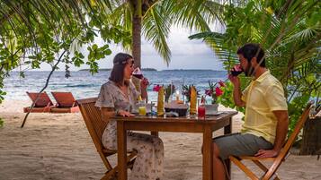 A wooden table with a candle, drinks, and a small vase of flowers on a beach.