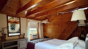 Un dormitorio con techo de madera, cama con ropa de cama blanca, mesita de noche con lámpara y ventana con cortinas.
