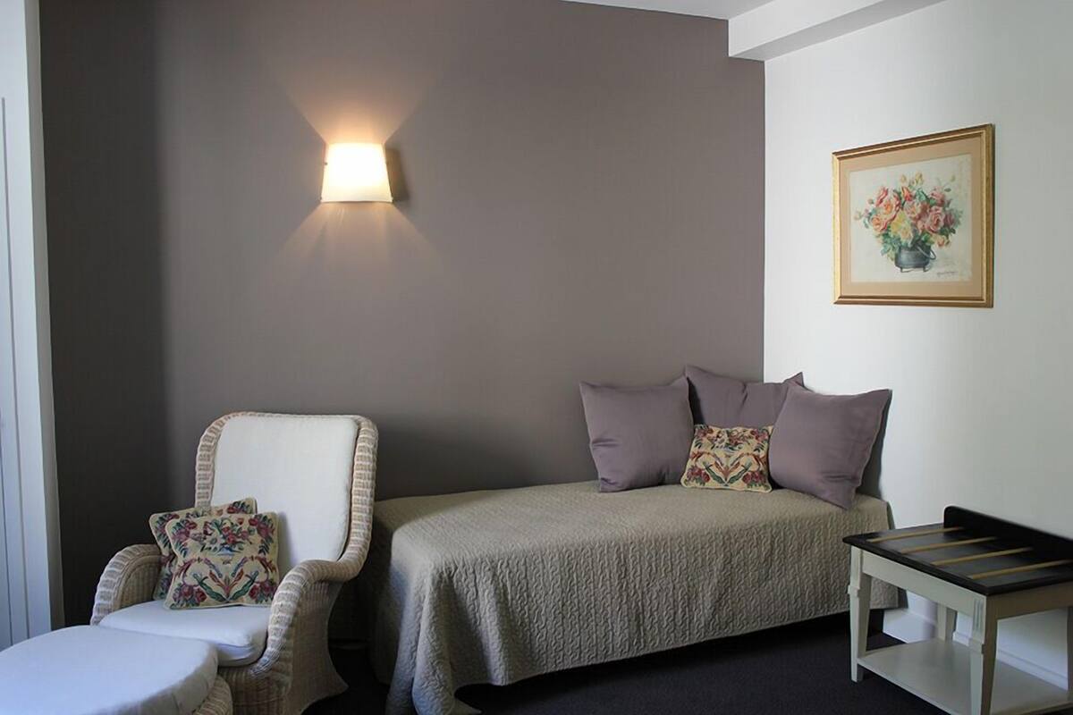 Una habitación con una cama, un sillón con cojín, una mesita y una lámpara fijada en la pared.