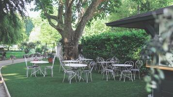 An outdoor seating area with white metal tables and chairs, surrounded by lush greenery and a large tree.