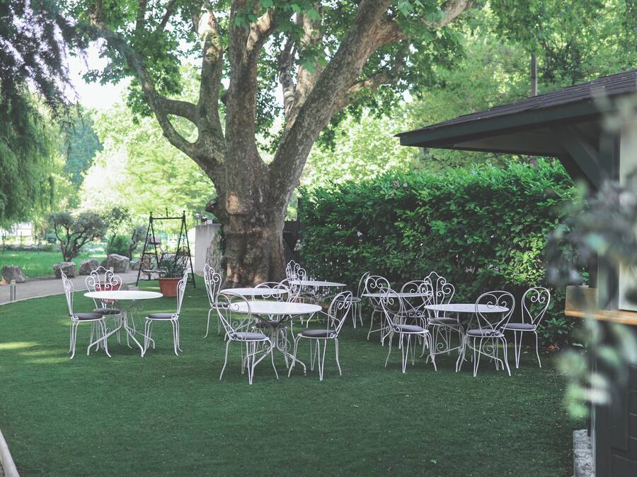 An outdoor seating area with white metal tables and chairs, surrounded by lush greenery and a large tree.