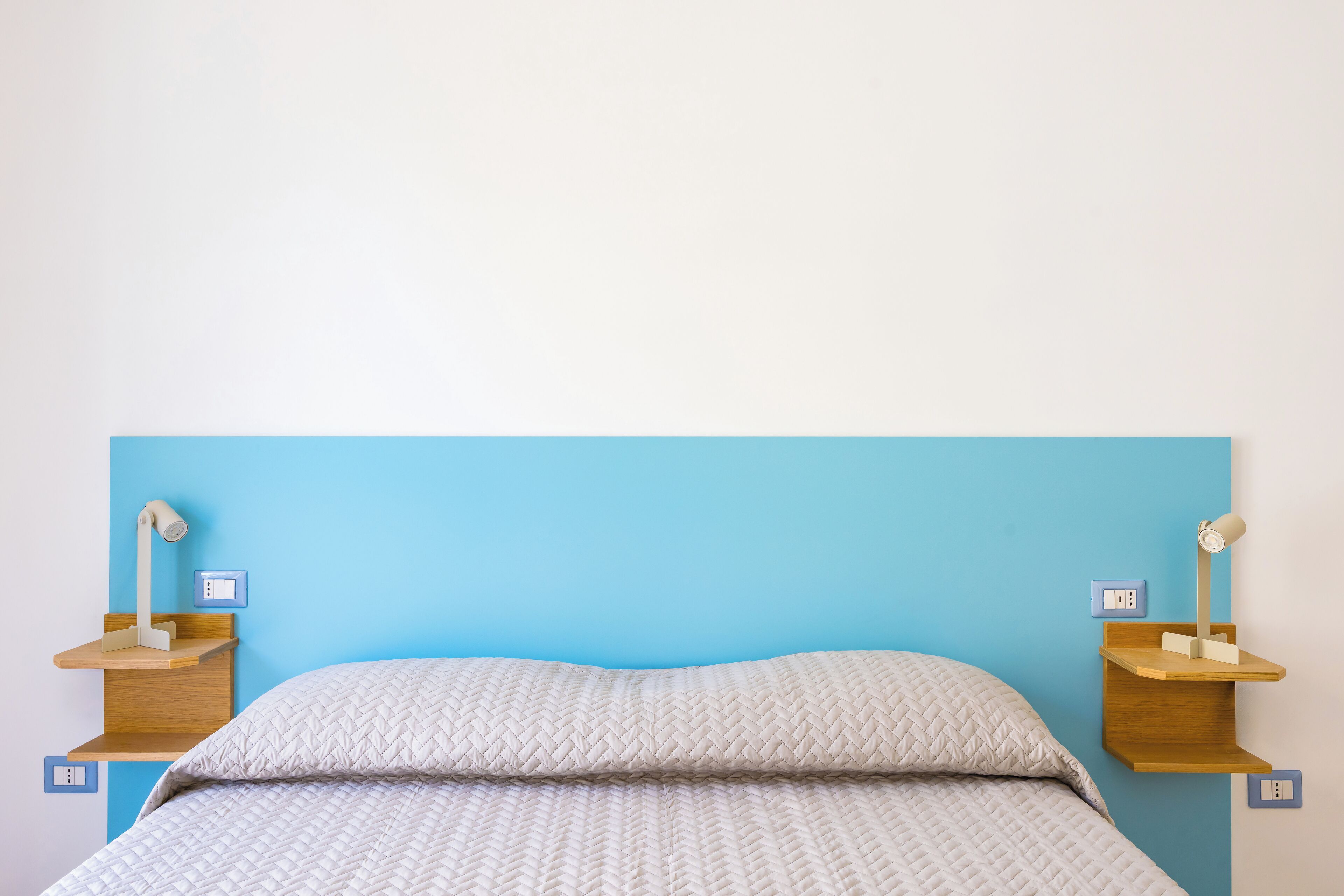 A bed with a quilted white duvet, two wooden bedside tables with lamps, and a blue wall.