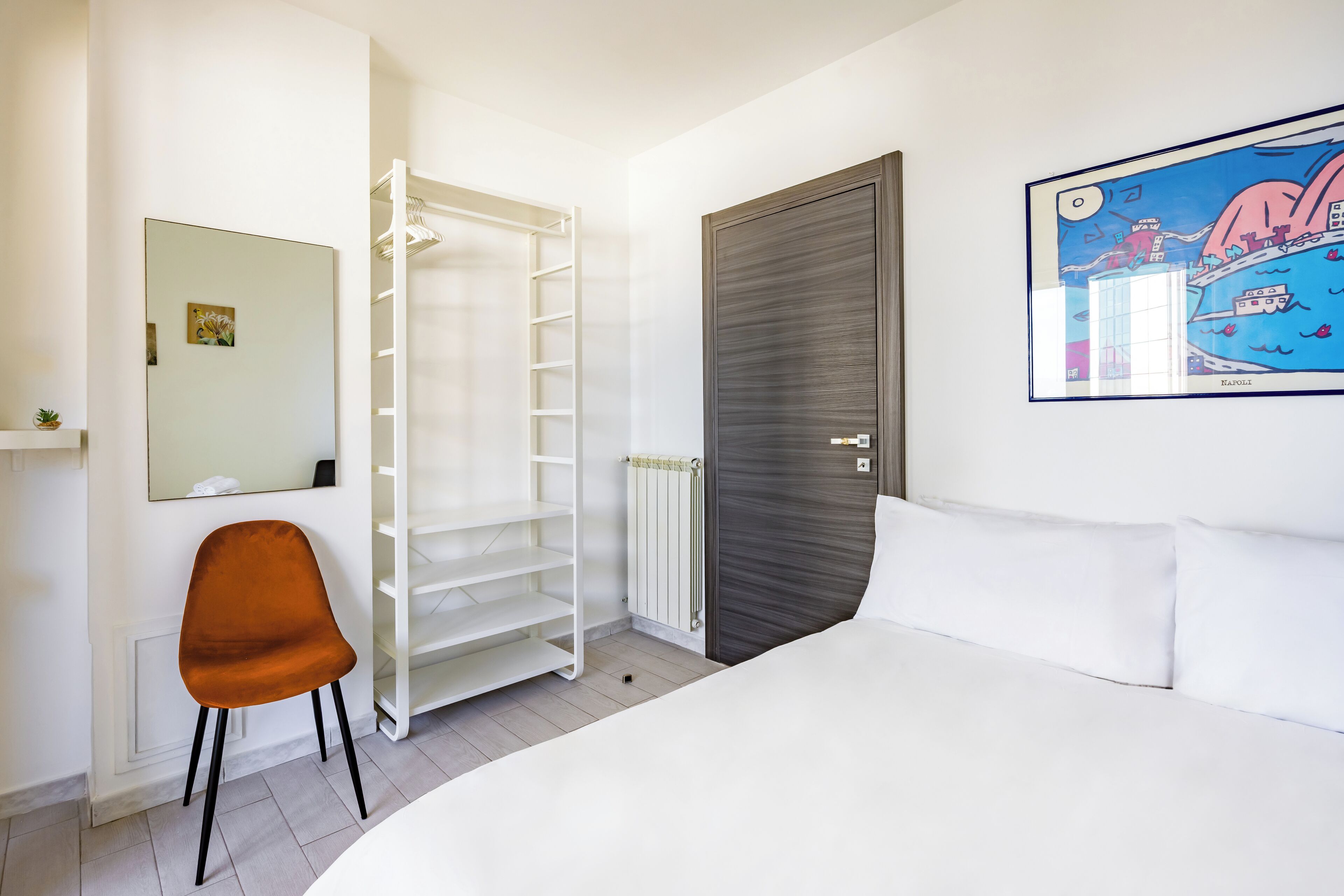A bedroom with a bed, a white chair, a wardrobe, a radiator, and a framed picture on the wall.