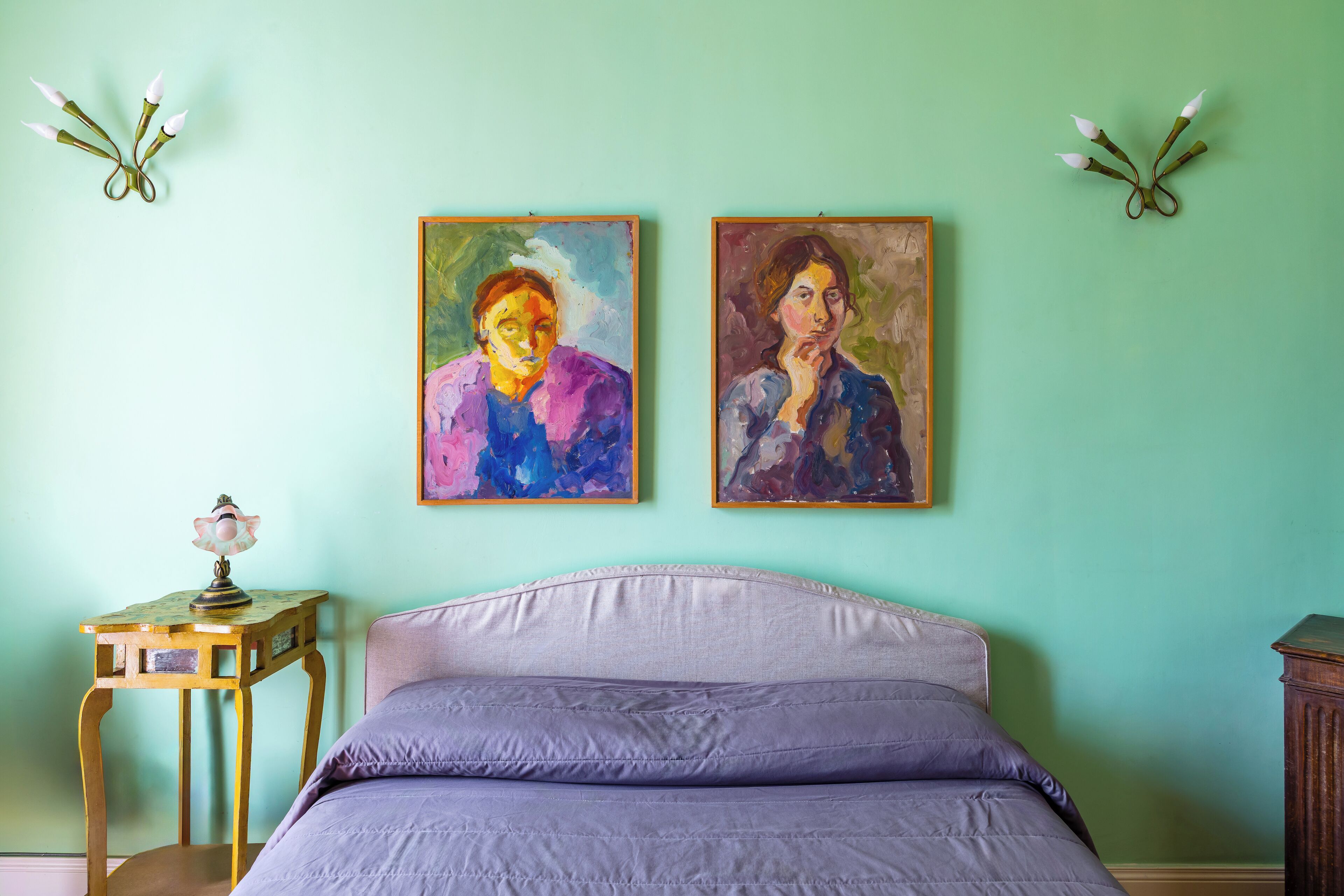 A bedroom with a bed, two paintings on the wall, a small table with a lamp, and a wooden nightstand.
