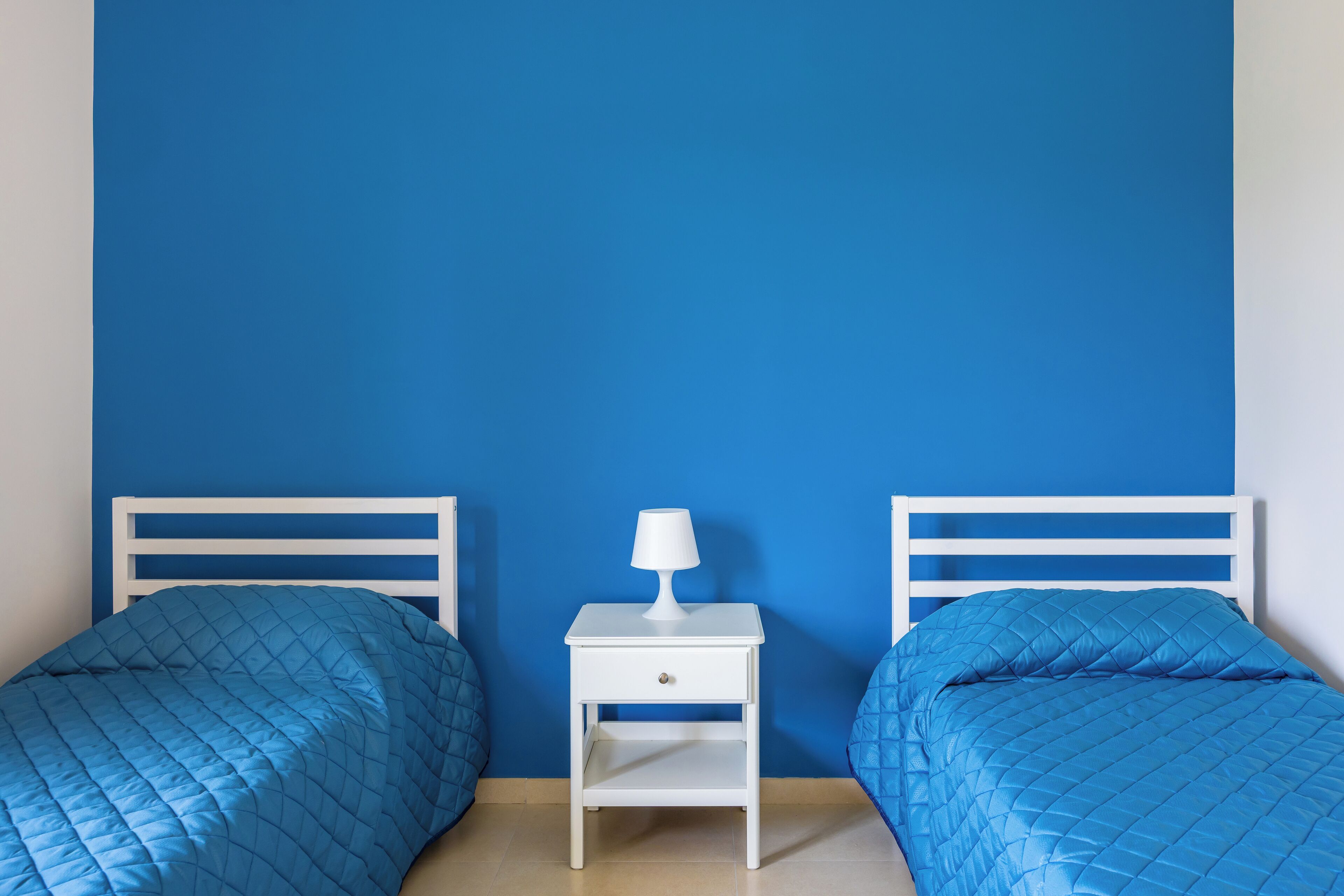 Ein Zimmer mit zwei Betten, jeweils mit einer blauen gesteppten Tagesdecke, einem weißen Nachttisch mit Lampe und einer blauen Wand.