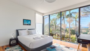 A modern bedroom with a large bed, a bedside table, and a view of palm trees and the ocean through floor-to-ceiling windows.