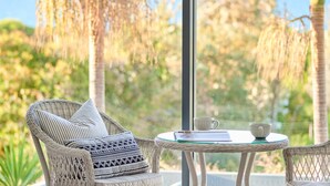 A wicker chair with patterned cushions, a glass table with a book and a cup, and a view of trees through a window.