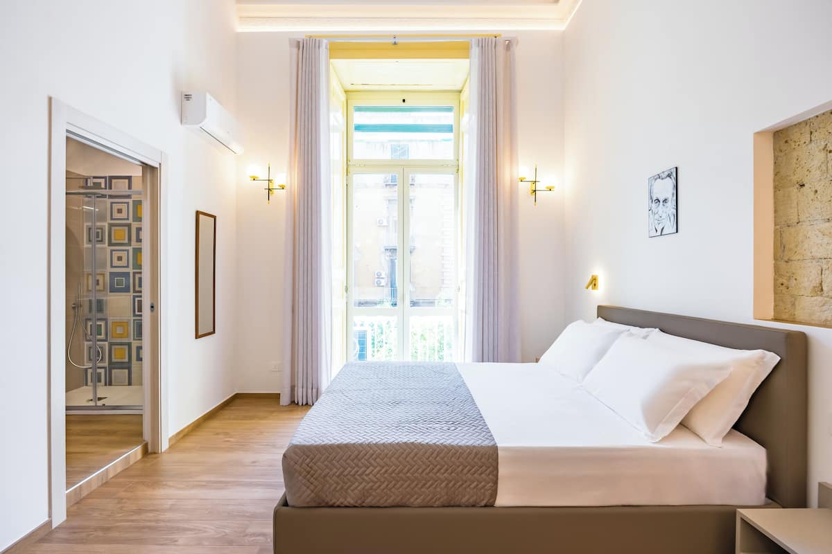 A hotel room with a bed, a window with curtains, a wall-mounted light, and a framed picture.