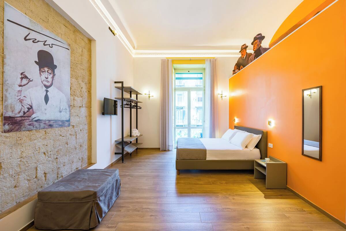 A hotel room with a bed, a bench, a TV, a mirror, and a poster on the wall.