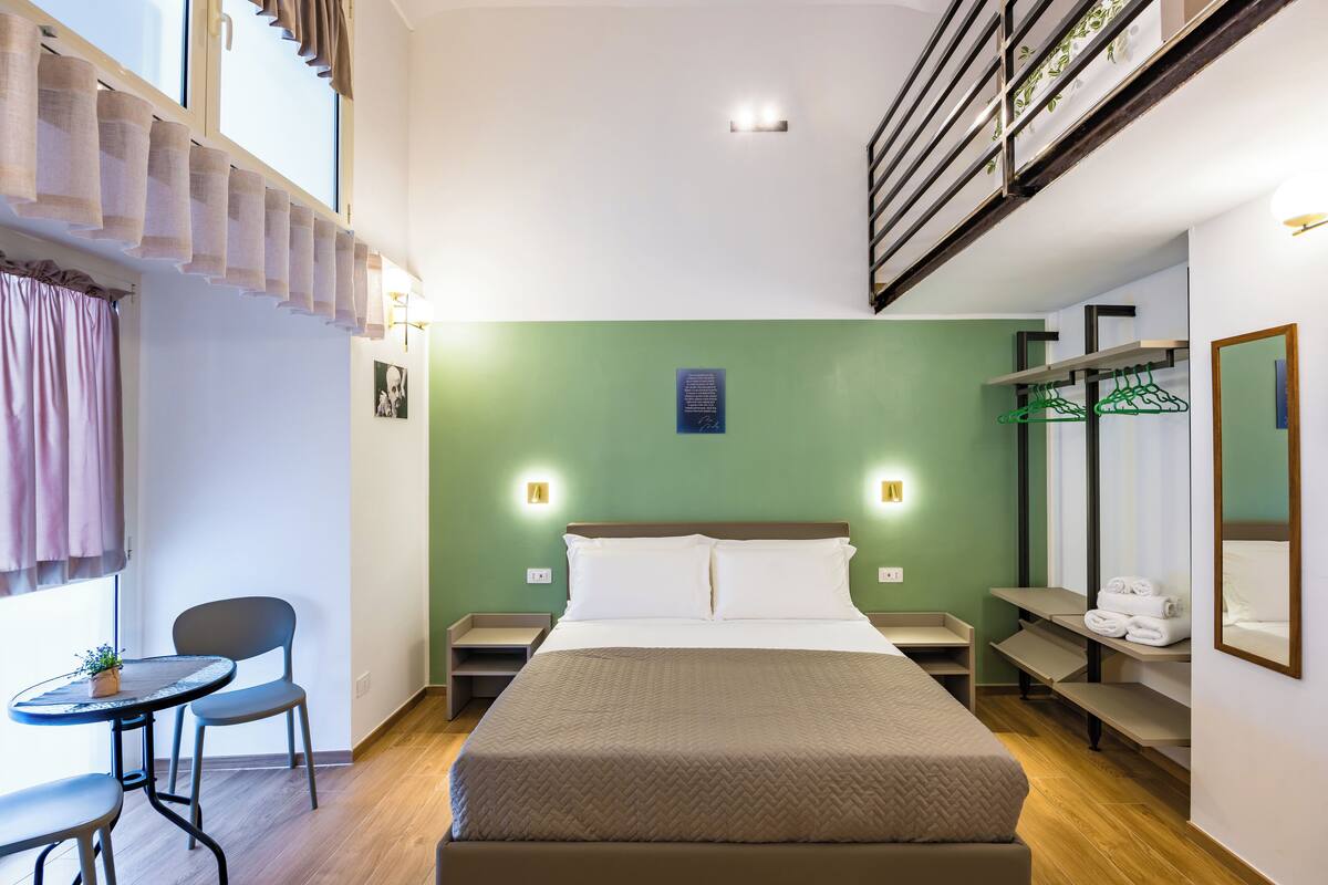 A hotel room with a large bed, a small table with chairs, a desk, and a hanging clothes rack.