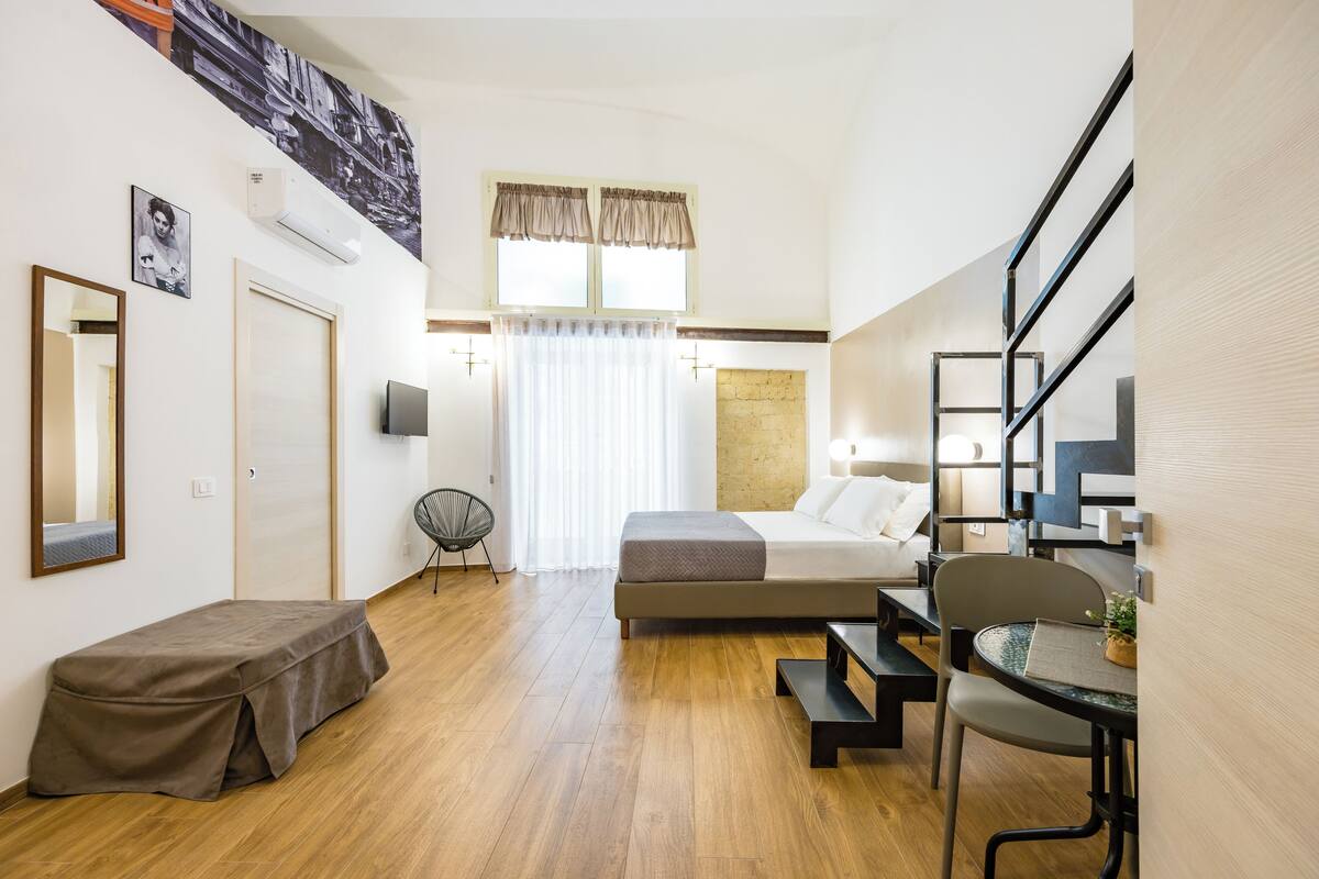 A modern hotel room with a bed, bedside table, chair, and a small desk with a chair. There is a window with curtains, a wall-mounted air conditioner, and a painting above the bed.
