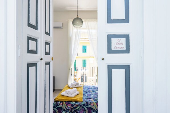 Un pasillo estrecho que conduce a una cama con colcha colorida, una mesita con toallas y un cartel en la puerta.