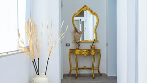 A hallway with a white wall, a gold-framed mirror, a cream-colored vase with decorative branches, and a gold-framed console table.