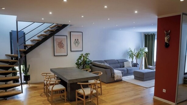 A spacious living area with a wooden staircase, a dining table with chairs, a grey sofa, and a red accent wall. - 3-bedroom house (Les Mages)