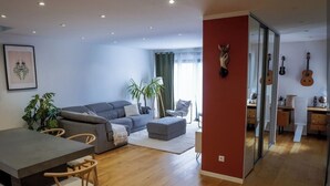 A modern living room with a grey sofa, a dining table with chairs, a small ottoman, and a kitchen area with cabinets and a guitar. - 3-bedroom house (Les Mages)
