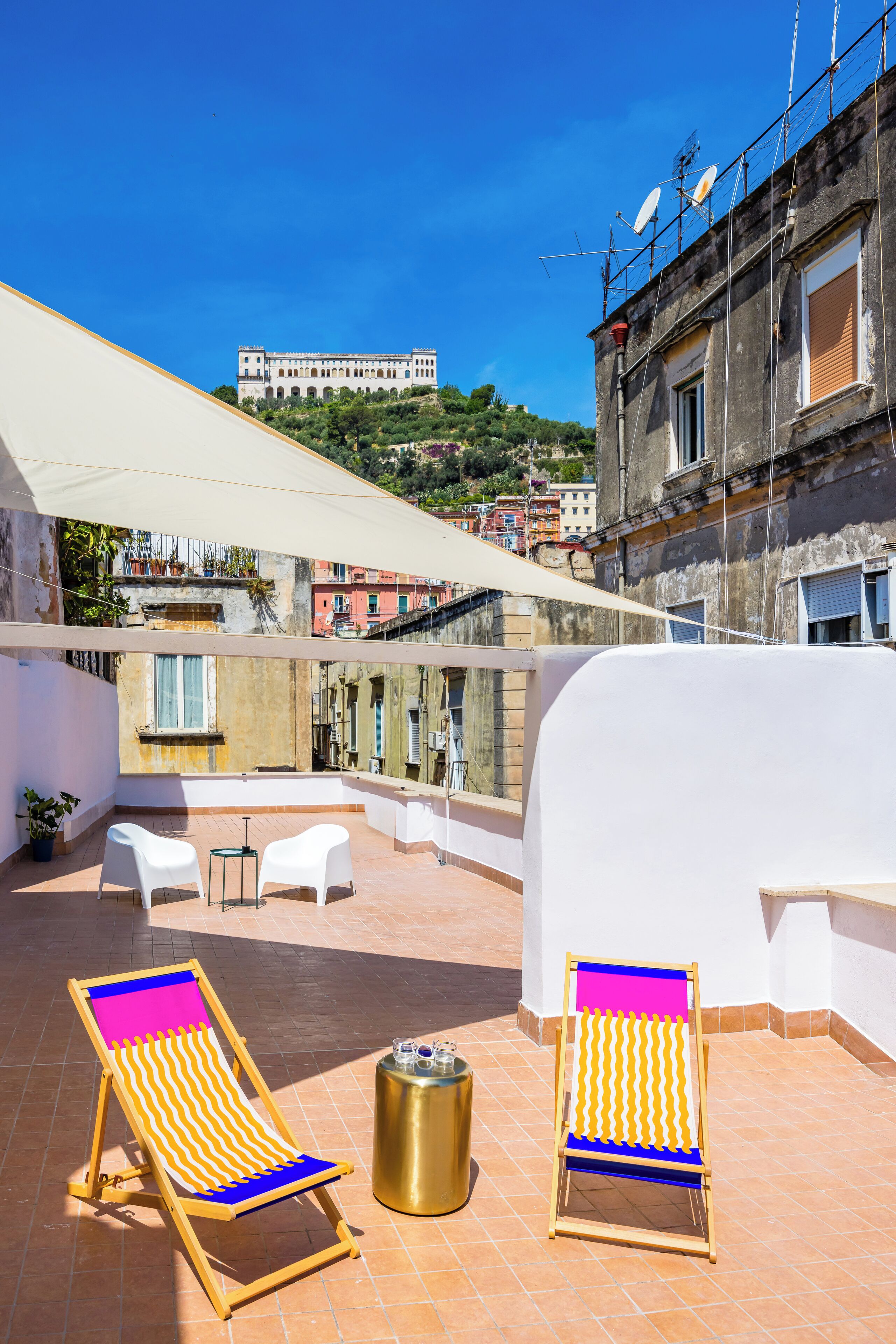 Eine Dachterrasse mit zwei gestreiften Liegestühlen, einem kleinen runden Tisch und einer goldenen Mülltonne. Die Terrasse bietet Blick auf eine Stadtlandschaft mit historischen Gebäuden und einer Hügelkulisse im Hintergrund.