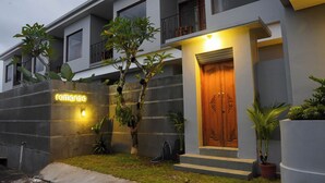 A modern building with a neon sign reading "romanso" on the facade, a wooden door with decorative carvings, and potted plants around the entrance. - Romansa Canggu (Canggu)