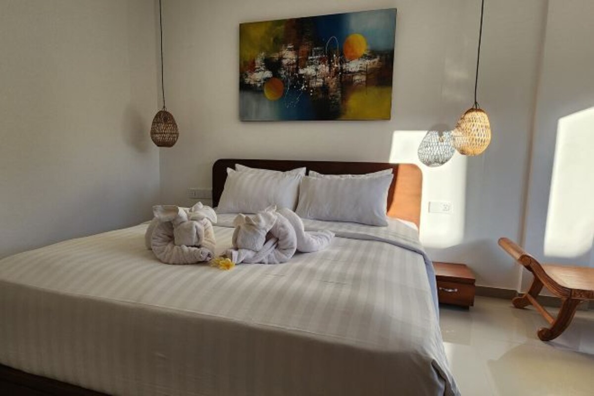 A neatly made bed with white linens and towels, a modern abstract painting on the wall, and a wooden chair in the corner.