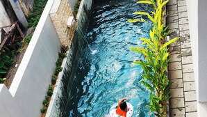 Ein Schwimmbecken mit einer Person in einem Ring, umgeben von Holzmöbeln und Pflanzen.