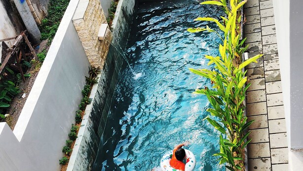 A swimming pool with a person in a ring, surrounded by wooden furniture and plants. - Romansa Canggu (Canggu)
