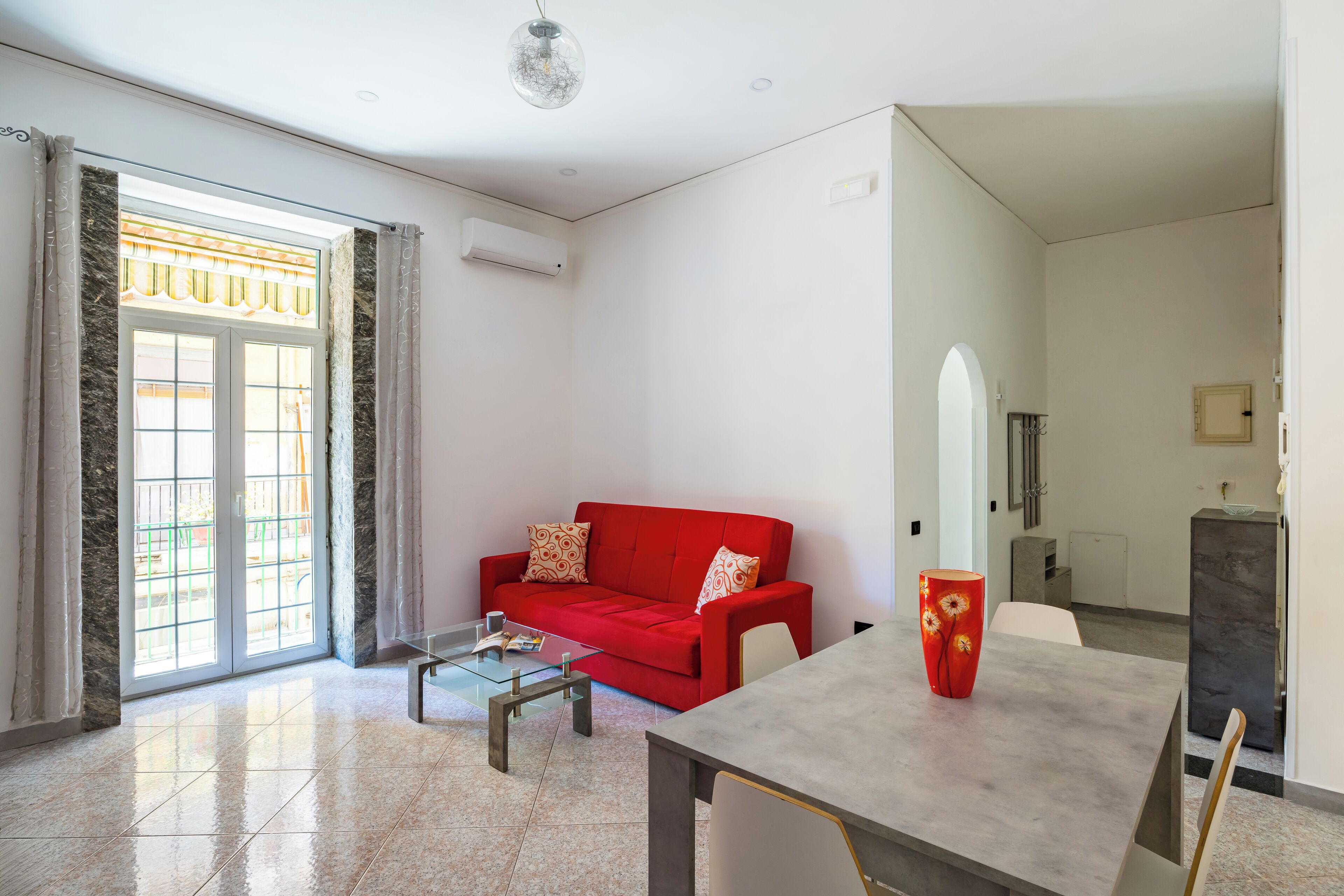 Una sala de estar con un sofá rojo, una mesa de centro de vidrio y una mesa de comedor con un jarrón.