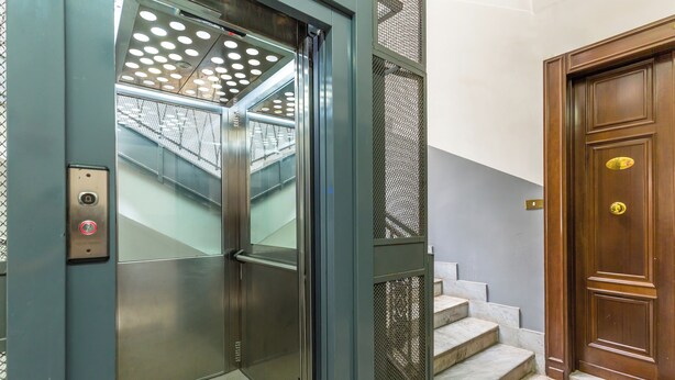 Un ascenseur doté d’une porte en métal et d’un panneau de commande, un escalier avec des marches en marbre et une porte en bois avec une poignée en laiton.