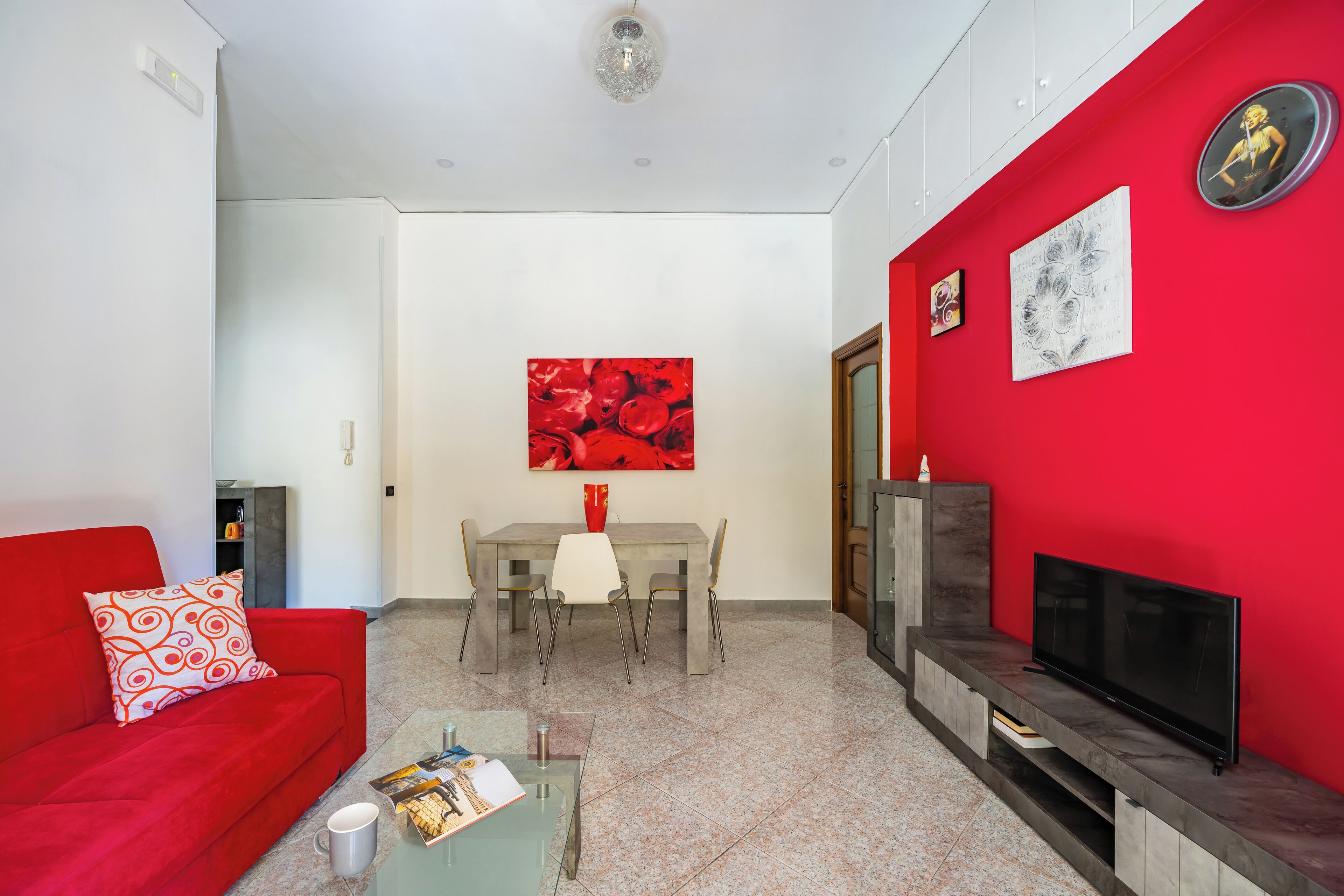 Un salon avec un canapé rouge, une table à manger entourée de chaises, un téléviseur posé sur un meuble et des œuvres d’art accrochées aux murs.