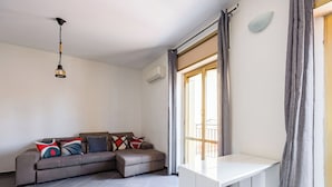 A modern living room with a sectional sofa, a hanging light fixture, a white desk, and a large window with curtains. - Revolutionary Apartment Near Duomo Station (Naples)