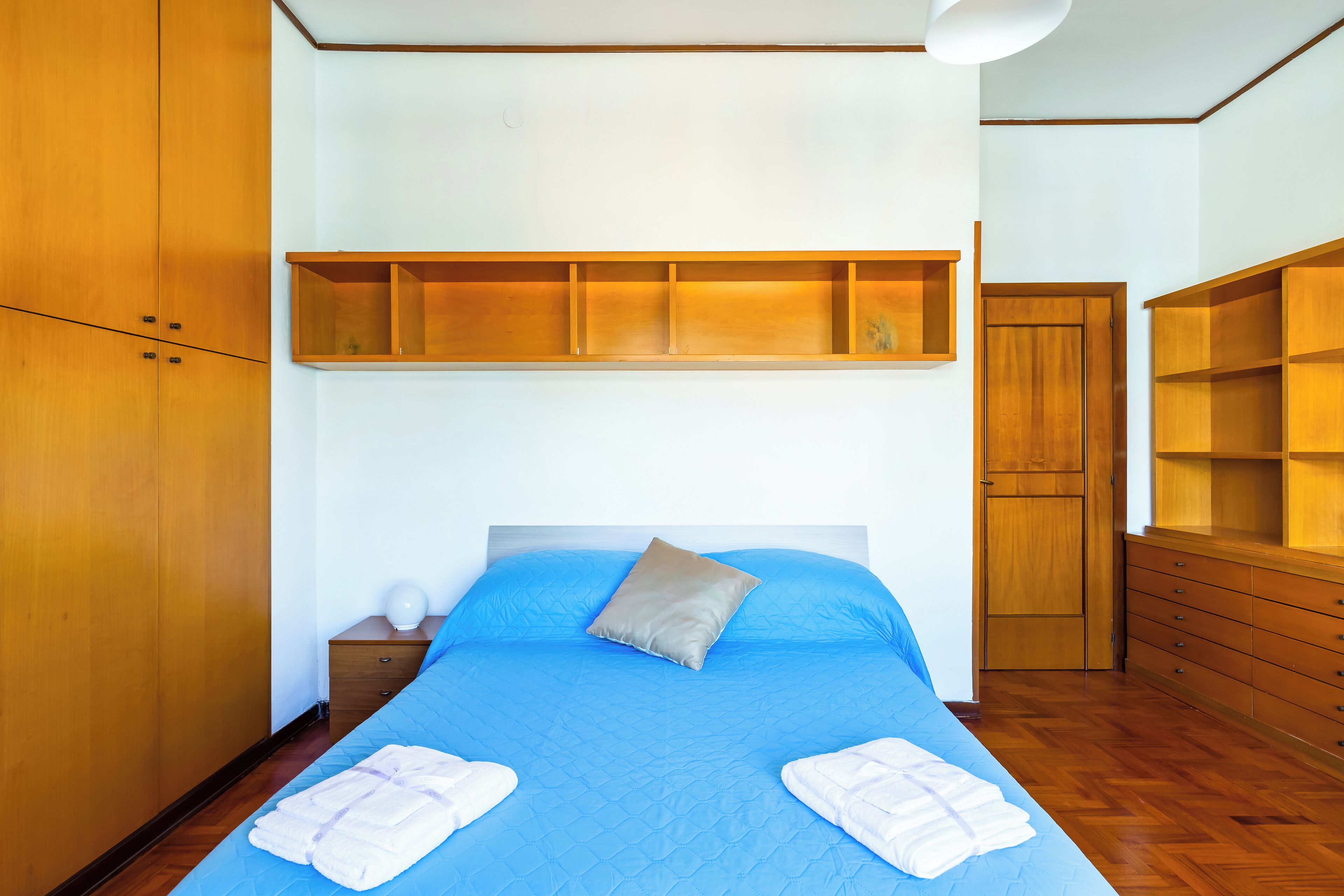 A bedroom with a blue bedspread, a wooden wardrobe, and a built-in shelf above the bed.