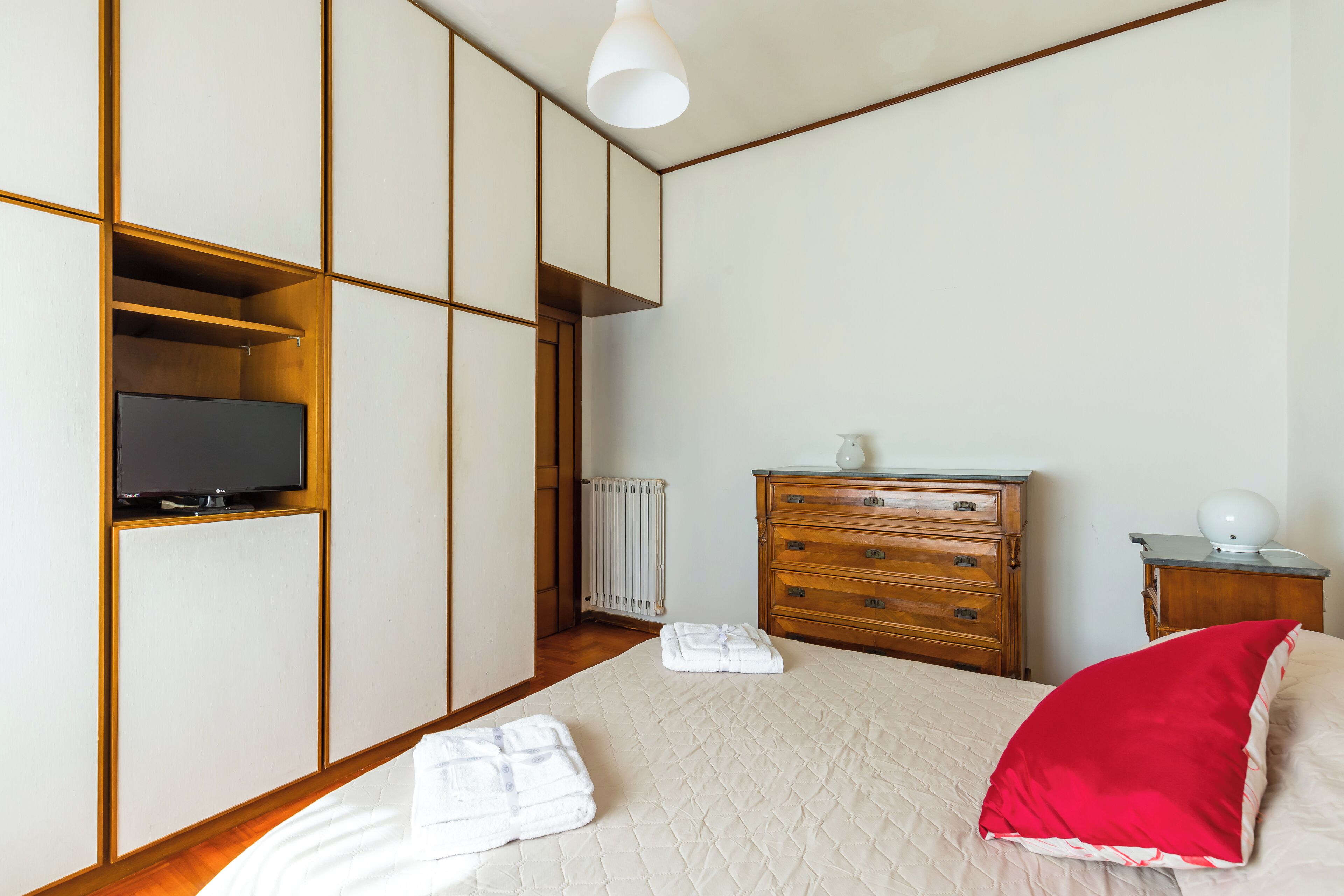Ein Schlafzimmer mit Bett, Holzkommode, Nachttisch, Fernseher und Heizkörper.