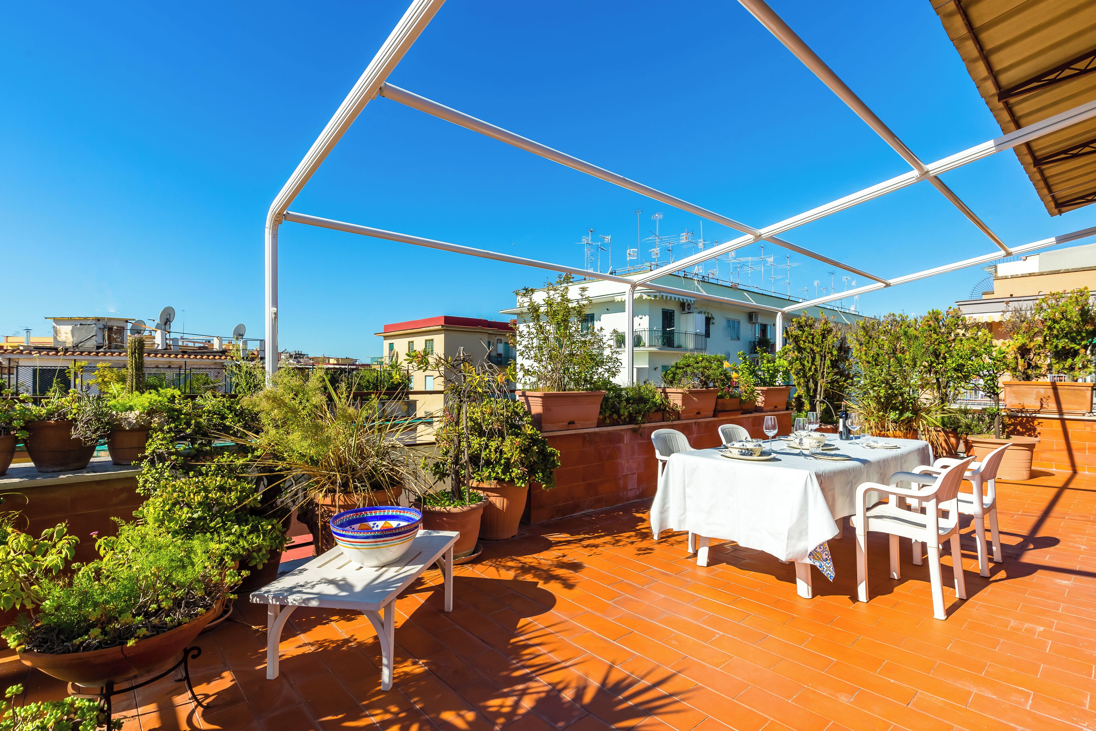 A rooftop terrace with a table set for dining, surrounded by potted plants and a clear blue sky.
