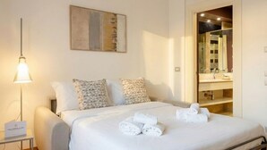 A hotel room with a bed, pillows, a lamp, a painting, and a bathroom visible through an open door.