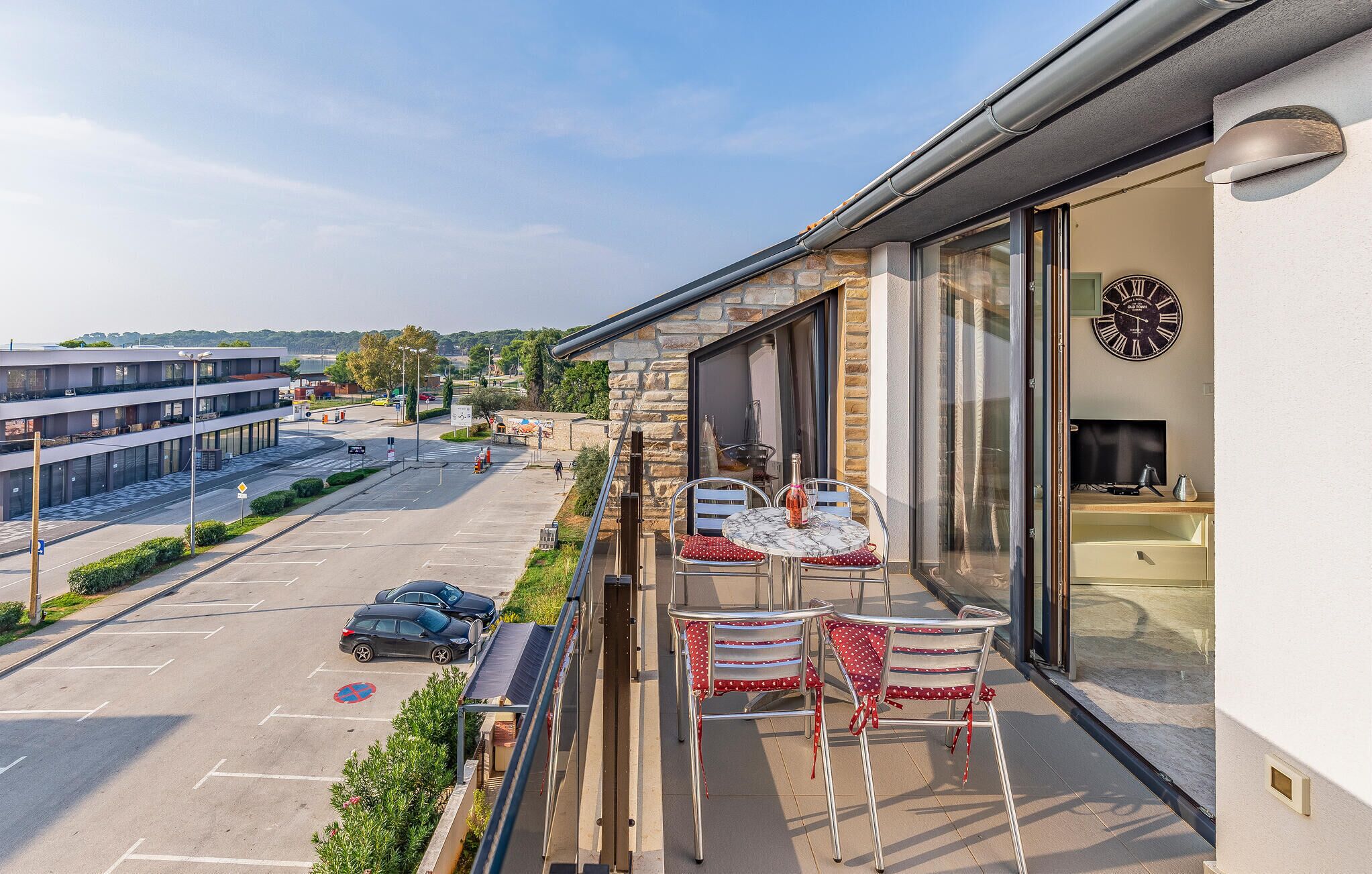Ein Balkon mit zwei Tischen, Stühlen, einer Uhr und Blick auf einen Parkplatz und Gebäude.