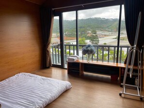 A hotel room with a bed, a small table with a vase of flowers, and a view of a hilly landscape through large windows. - DA LAT THIEN VUONG HOMESTAY (Lac Duong)