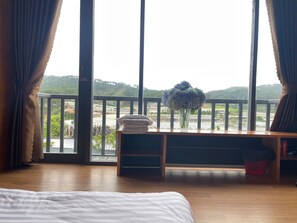 A bedroom with a large window, a bed, a bedside table with a vase of flowers, and a view of a landscape. - DA LAT THIEN VUONG HOMESTAY (Lac Duong)