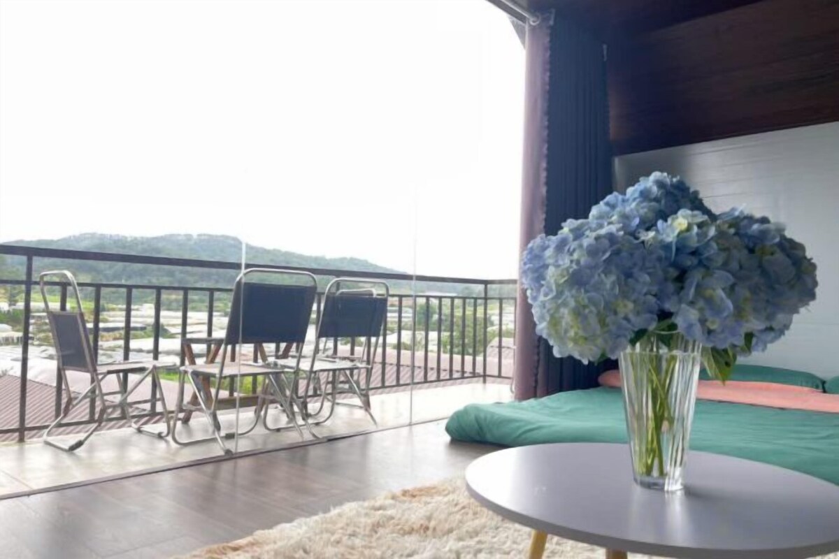 A balcony with a view of a river and hills, a table with a vase of flowers, and a bed with a green pillow.