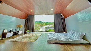 Un dormitorio con una cama grande, un escritorio pequeño y vistas a un paisaje verde a través de un balcón.