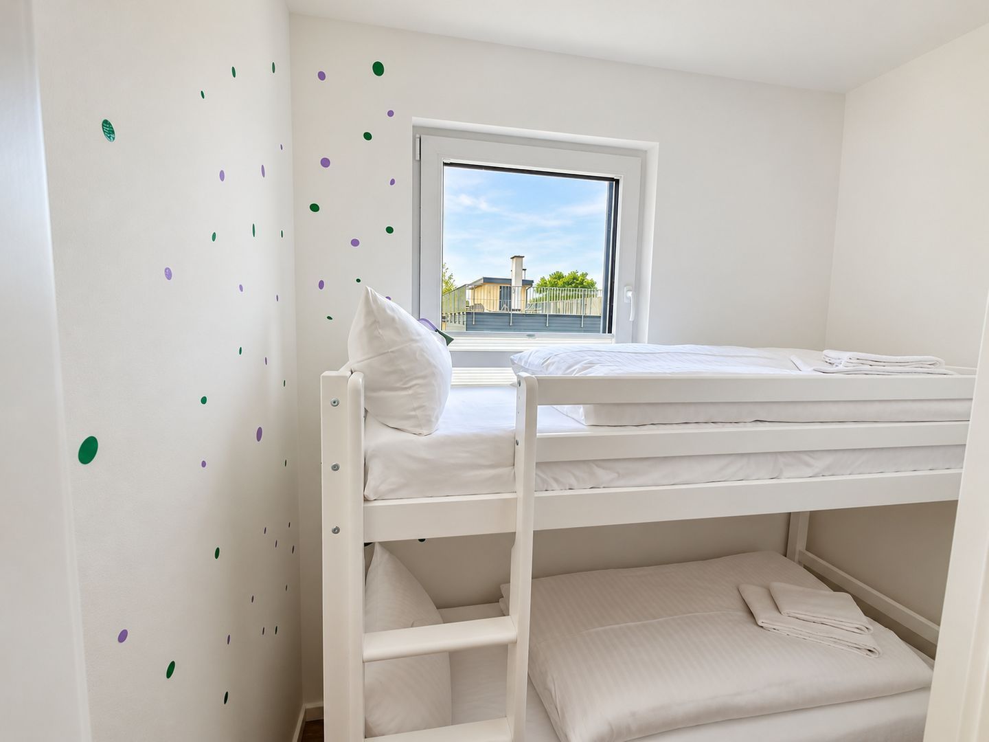 Camera con letto a castello, finestra, pareti bianche e vista sugli edifici.