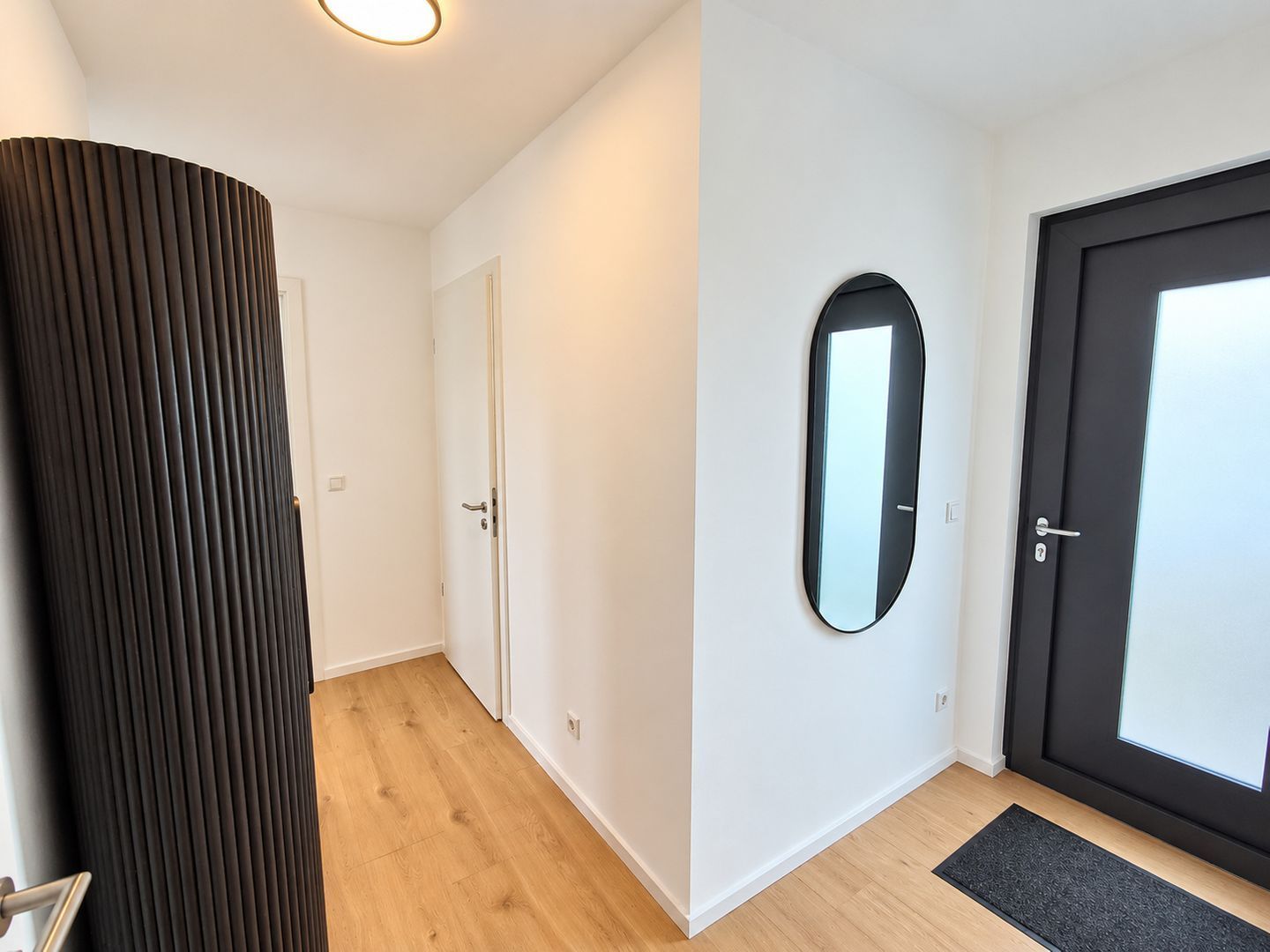 A hallway with a wooden floor, a black door, a black-framed mirror, and a white wall.