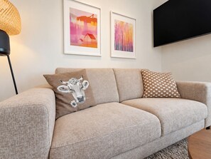 A modern living room with a beige sofa, two framed artworks, a decorative pillow with a cow design, and a black TV on the wall.