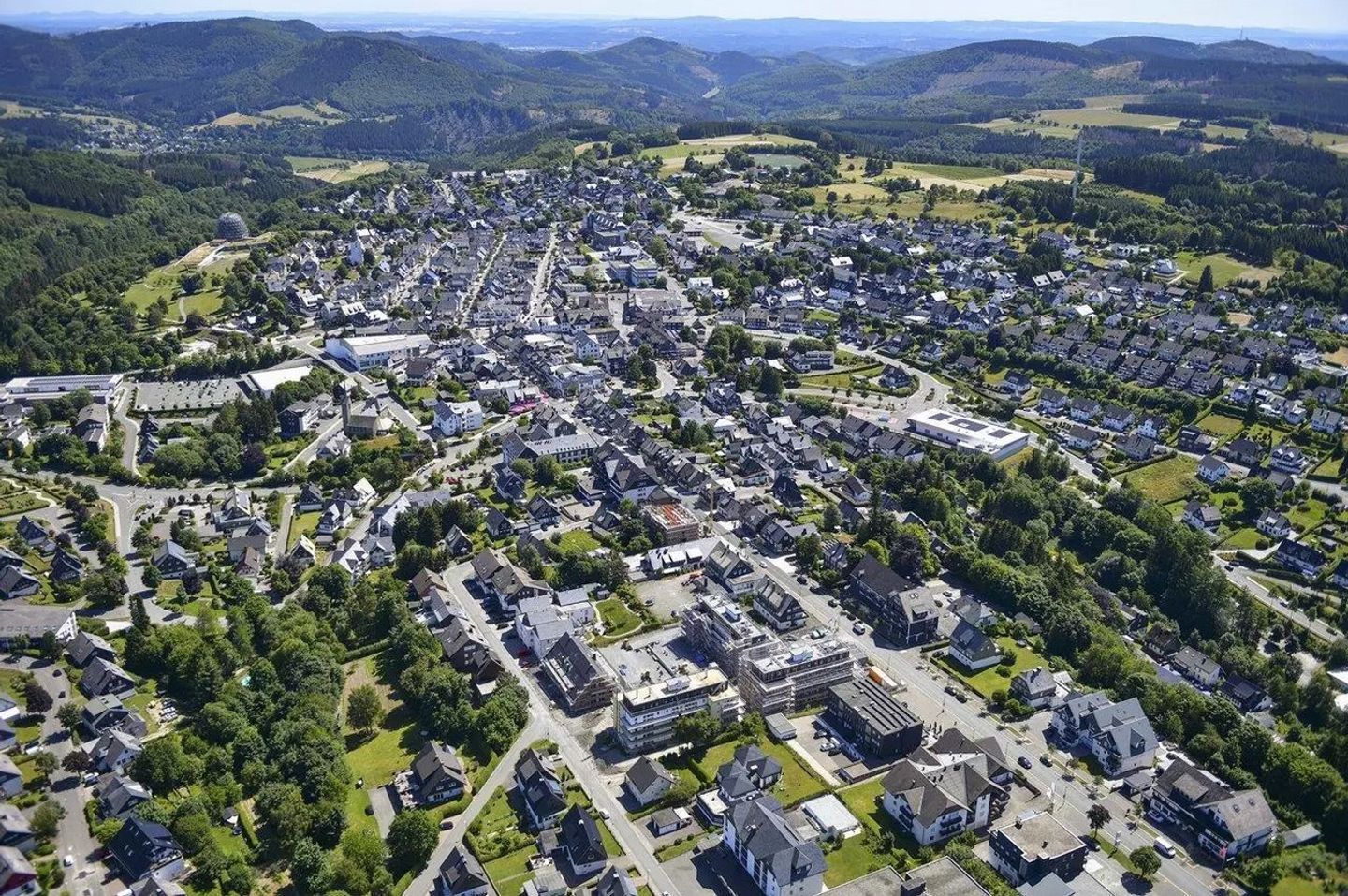 Une vue aérienne d’une ville avec des maisons d’habitation, un grand bâtiment surmonté d’un dôme et des collines verdoyantes environnantes.