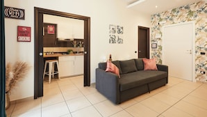 A modern living room with a grey sofa, a kitchen area with a counter and stools, and a door with a tropical wallpaper. - Vicky's Luxury Guesthouse by Napoliapartments (Naples)