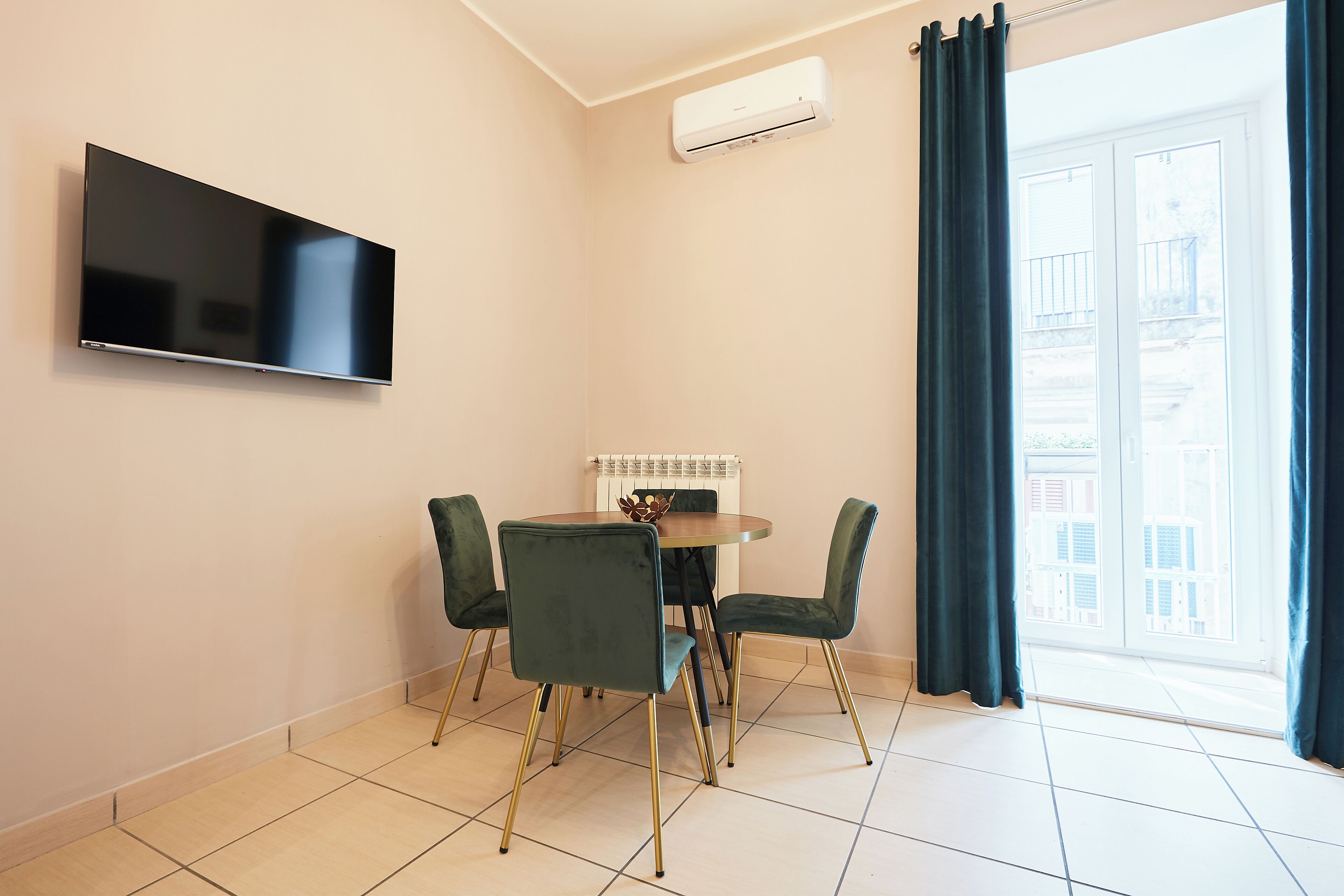 A room with a flat-screen TV mounted on the wall, a round table with four chairs, an air conditioning unit, and a view of buildings through a large window with curtains.