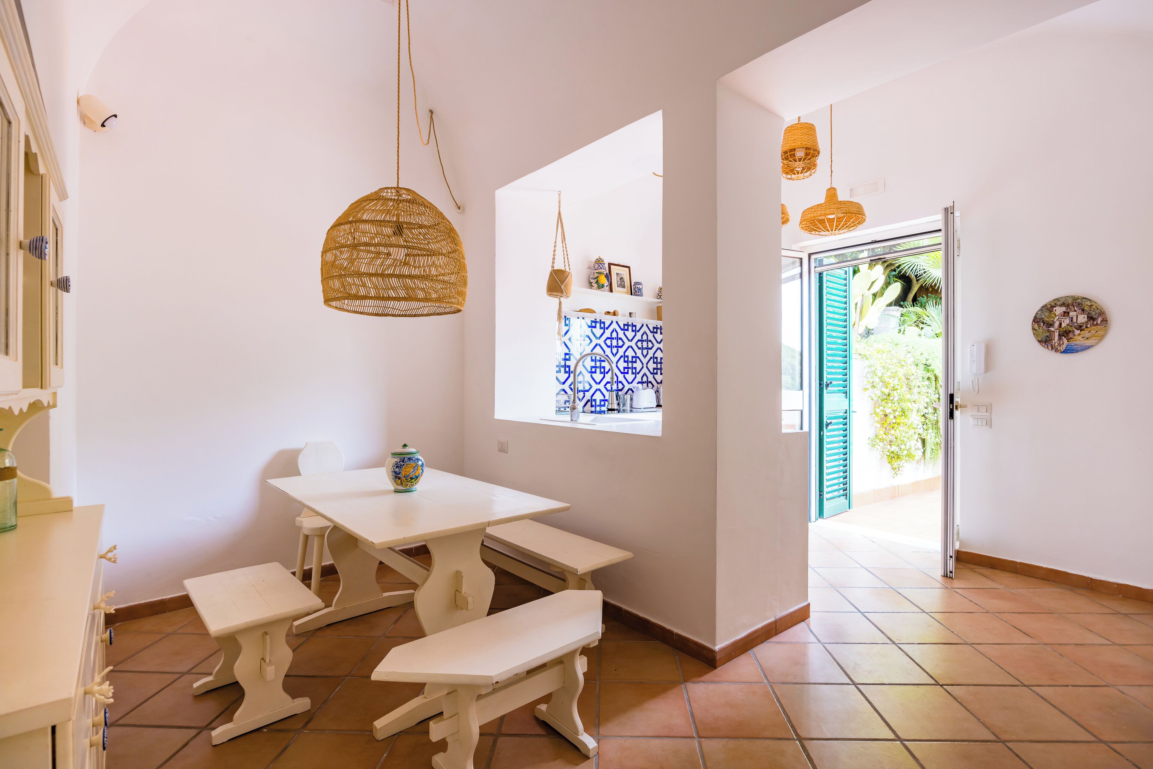 Um espaço amplo e iluminado com uma mesa branca e bancos, uma luminária de vime pendente e uma janela com cortina estampada em azul e branco.