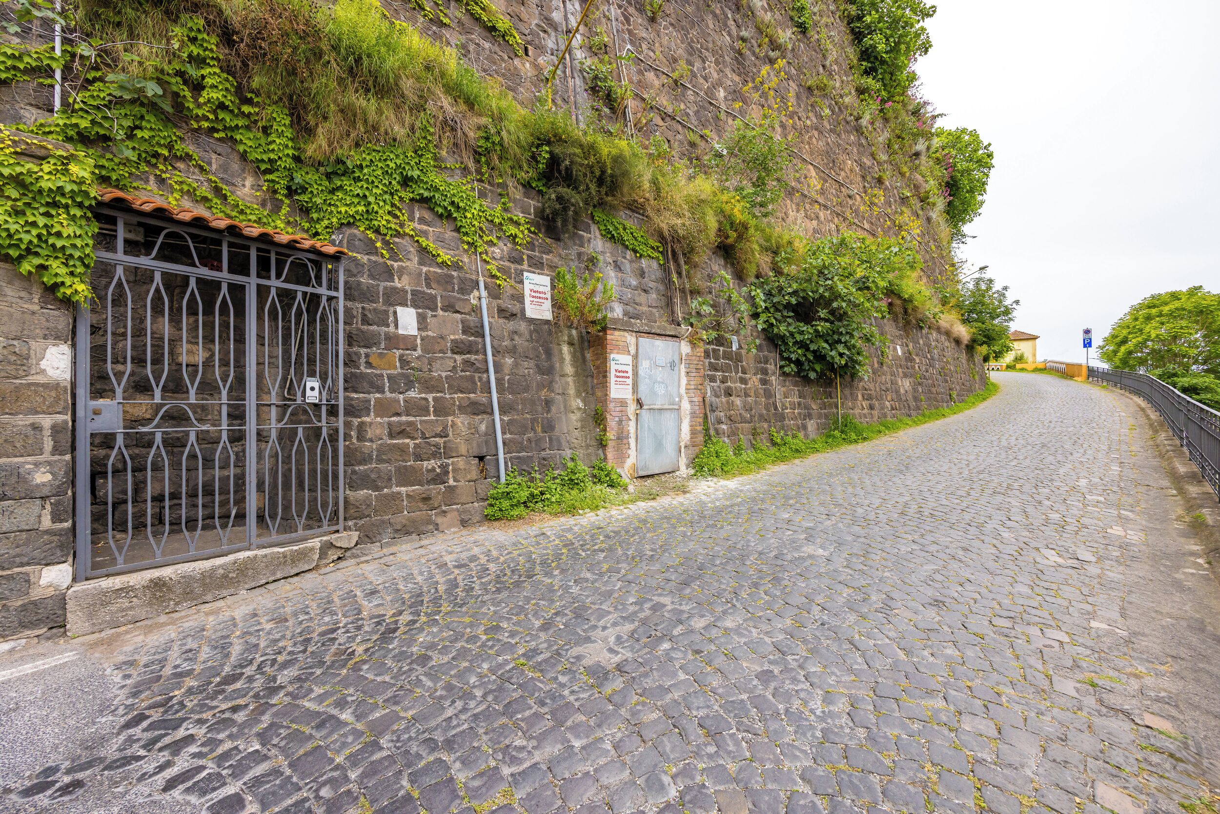 A cobblestone street with a stone wall on one side, a metal gate, and a small building with a blue door.