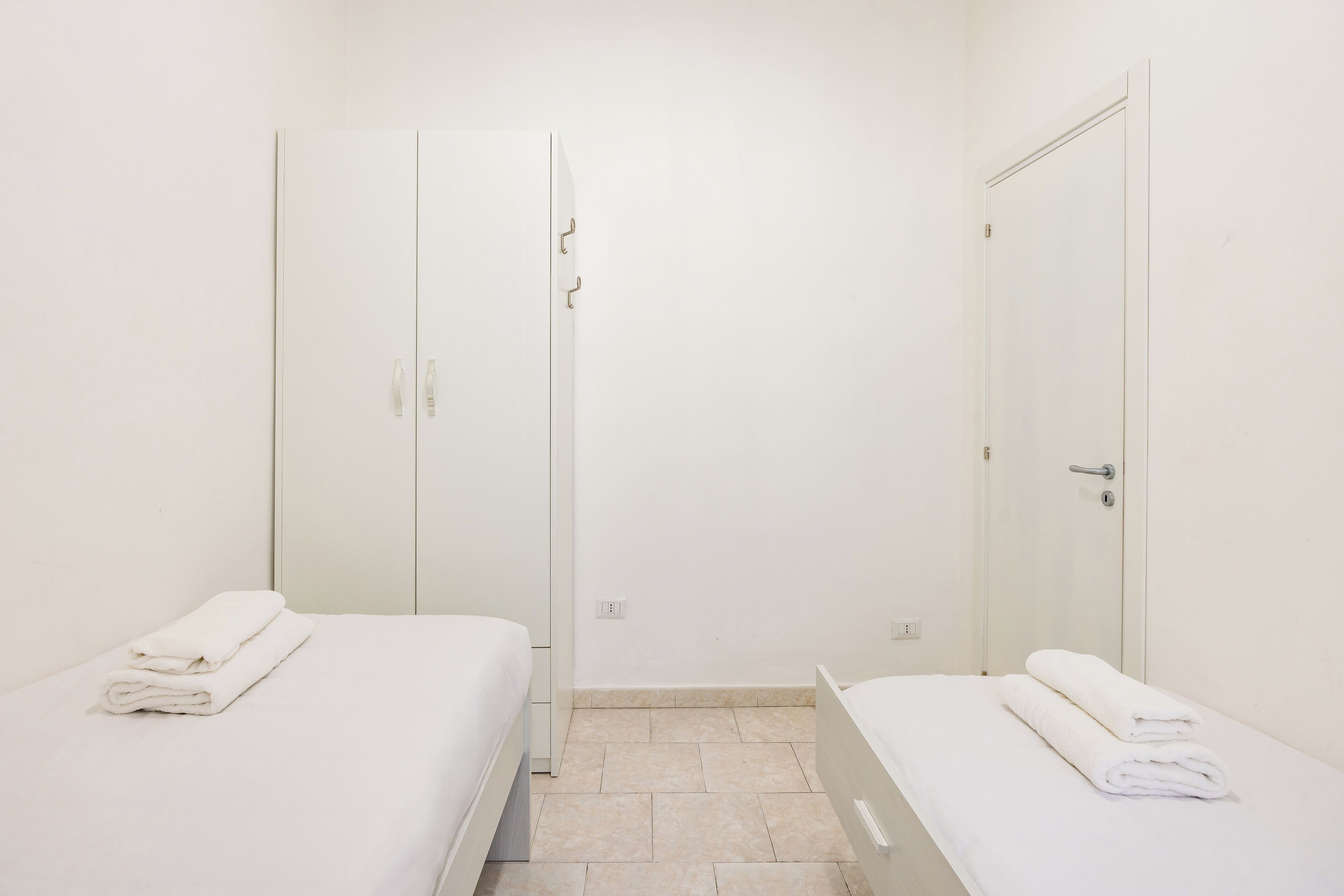 Ein Zimmer mit zwei Betten, jeweils mit einem ordentlich gefalteten weißen Handtuch. Weiße Schränke mit Griffen und eine Tür mit silbernem Griff sind ebenfalls vorhanden.