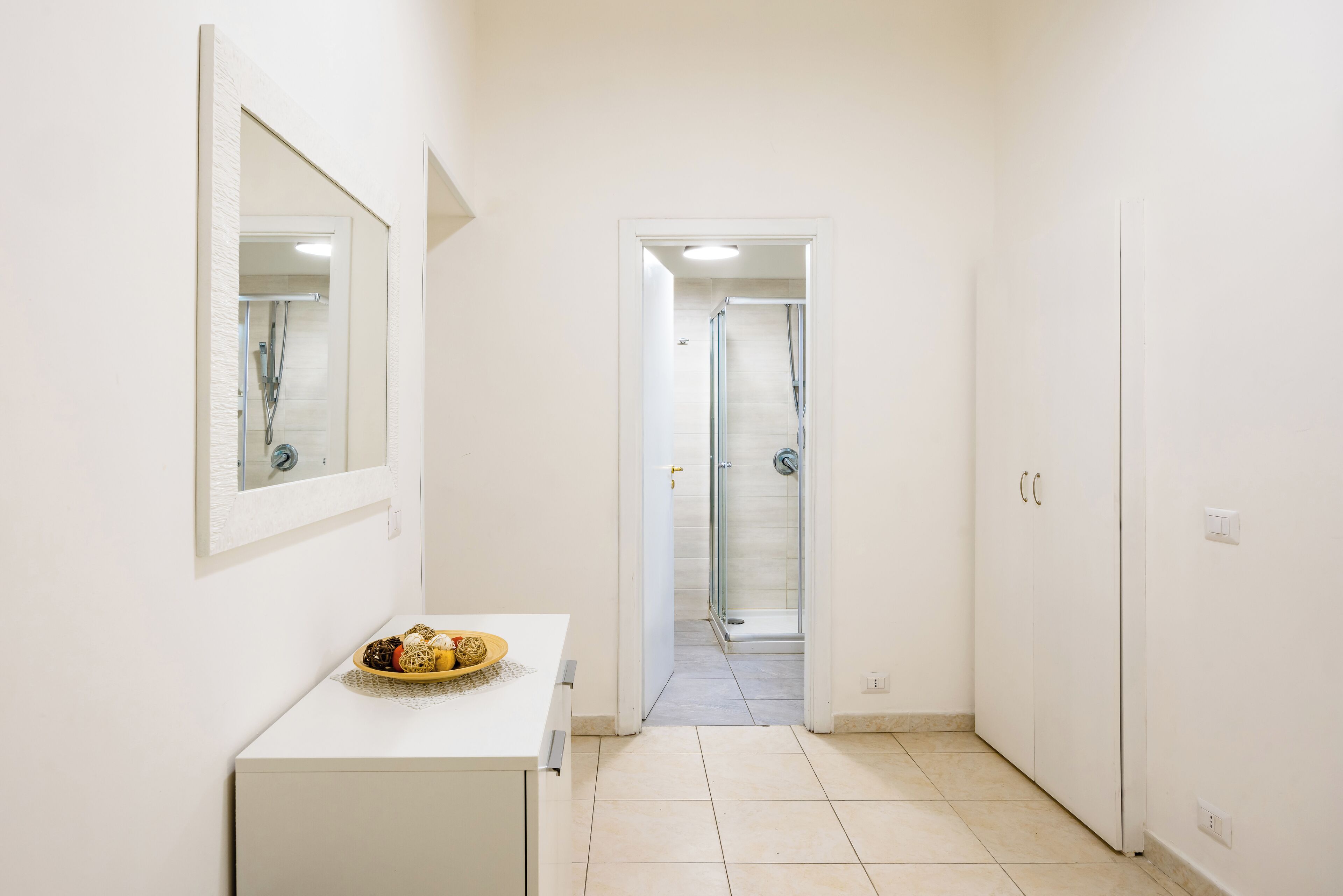 A narrow hallway with a white sink and mirror on the left, leading to a glass shower door.